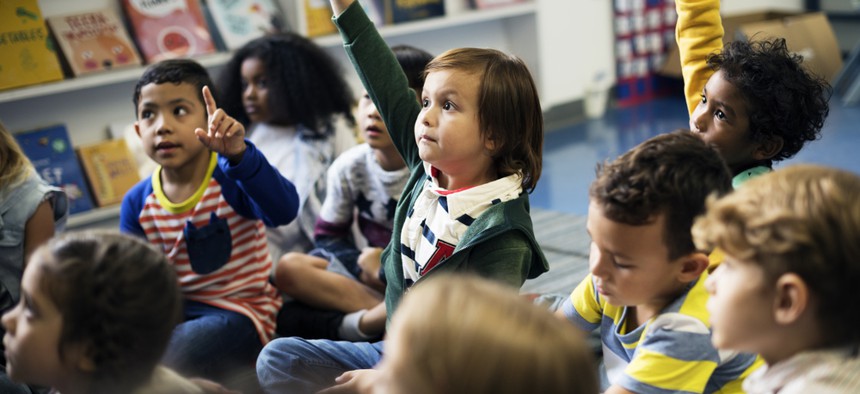 A child holds her hand up in a classroom with other children.