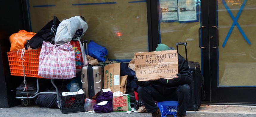 Homeless man sits on the street with a sign and belongings stacked on a shopping cart. 